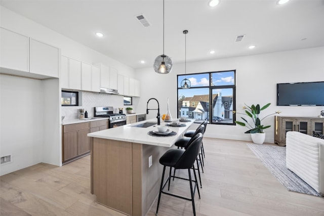 kitchen featuring stainless steel range with gas cooktop, decorative light fixtures, an island with sink, sink, and white cabinets