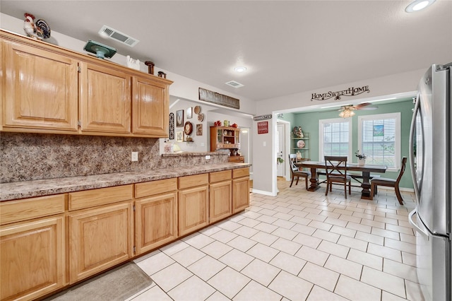 kitchen with light brown cabinetry, stainless steel refrigerator, backsplash, light tile patterned floors, and light stone counters