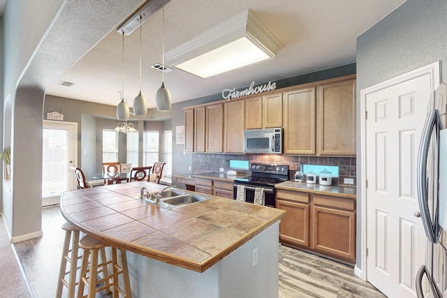 kitchen with sink, hanging light fixtures, appliances with stainless steel finishes, a kitchen island with sink, and backsplash