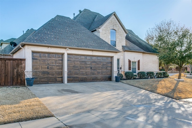 french country inspired facade with a garage, driveway, brick siding, and roof with shingles