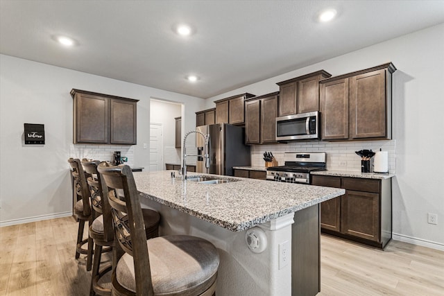 kitchen with appliances with stainless steel finishes, a breakfast bar area, a kitchen island with sink, light stone counters, and light wood-type flooring