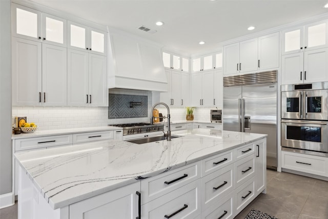 kitchen with stainless steel appliances, custom exhaust hood, light stone countertops, and a kitchen island with sink