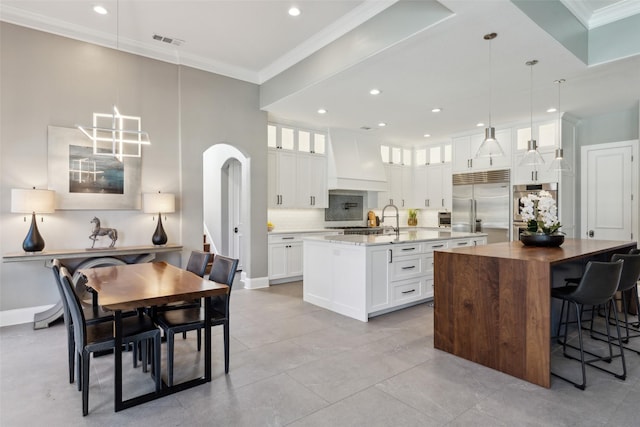 kitchen with pendant lighting, built in refrigerator, a kitchen island with sink, and custom exhaust hood