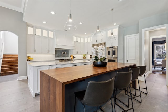 kitchen with butcher block countertops, a center island with sink, double oven, white cabinets, and backsplash