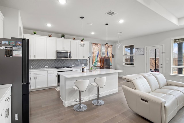 kitchen with a kitchen island with sink, hanging light fixtures, white cabinetry, backsplash, and stainless steel appliances