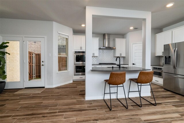 kitchen featuring a breakfast bar area, white cabinetry, stainless steel appliances, a kitchen island with sink, and wall chimney range hood