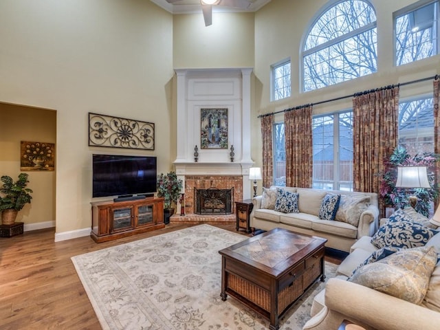 living room featuring a high ceiling, wood finished floors, a ceiling fan, baseboards, and a brick fireplace