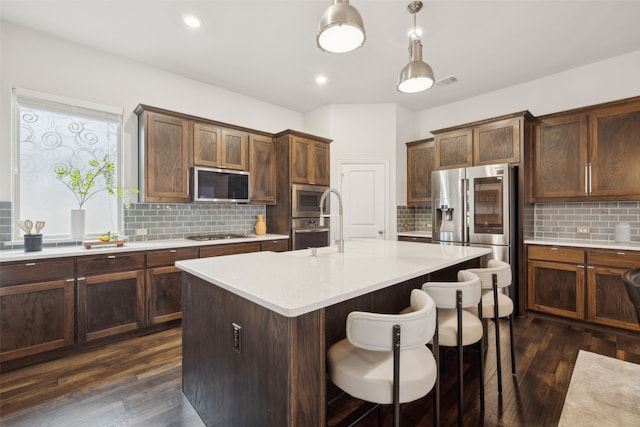 kitchen featuring dark wood-type flooring, an island with sink, pendant lighting, stainless steel appliances, and backsplash