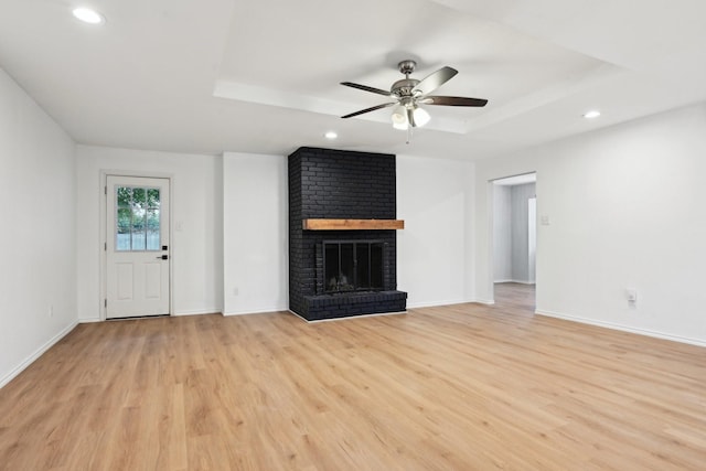 unfurnished living room featuring a raised ceiling, ceiling fan, a brick fireplace, and light hardwood / wood-style floors