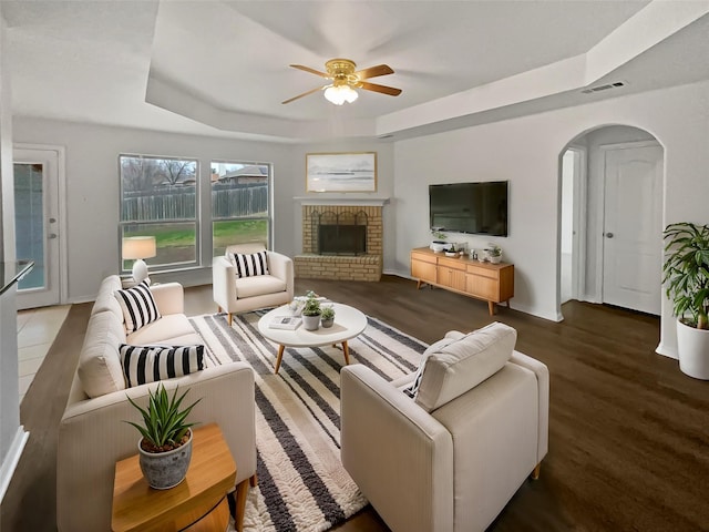 living room with a brick fireplace, dark hardwood / wood-style floors, a raised ceiling, and ceiling fan