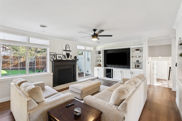 living room with built in features, wood-type flooring, ornamental molding, ceiling fan, and a brick fireplace