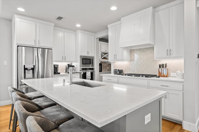 kitchen with white cabinetry, custom exhaust hood, stainless steel appliances, and an island with sink