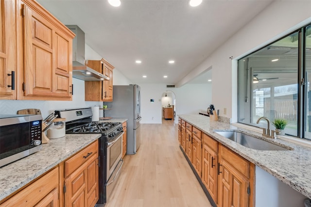 kitchen with wall chimney exhaust hood, sink, light stone counters, light wood-type flooring, and appliances with stainless steel finishes