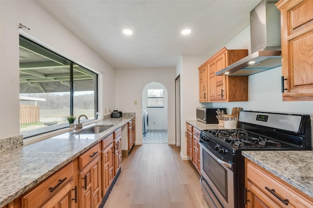 kitchen with wall chimney exhaust hood, sink, light wood-type flooring, stainless steel appliances, and light stone countertops