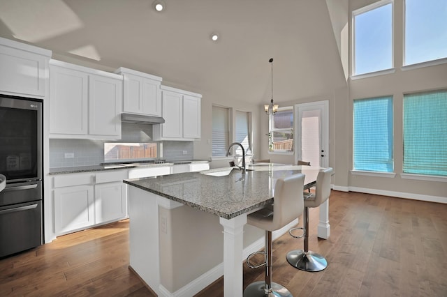 kitchen featuring sink, a center island with sink, hanging light fixtures, dark stone counters, and white cabinets