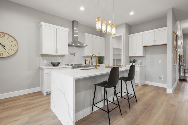 kitchen with white cabinets, a kitchen island with sink, light wood-type flooring, wall chimney range hood, and a sink