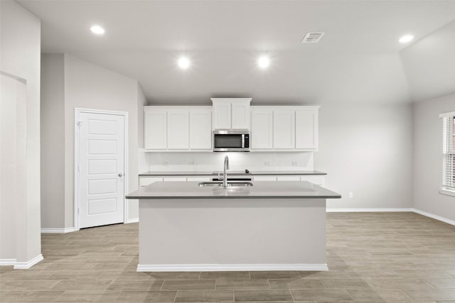 kitchen featuring visible vents, lofted ceiling, stainless steel microwave, a kitchen island with sink, and a sink