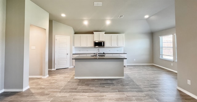 kitchen featuring lofted ceiling, stainless steel microwave, visible vents, a sink, and an island with sink