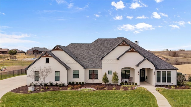 french provincial home featuring roof with shingles, a chimney, stucco siding, fence, and a front lawn