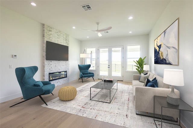 living room with ceiling fan, a stone fireplace, light hardwood / wood-style floors, and french doors