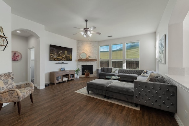 living room featuring dark wood-type flooring, ceiling fan, and a stone fireplace