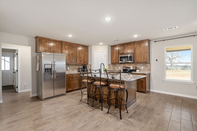 kitchen featuring appliances with stainless steel finishes, a kitchen breakfast bar, a kitchen island with sink, light stone counters, and a healthy amount of sunlight