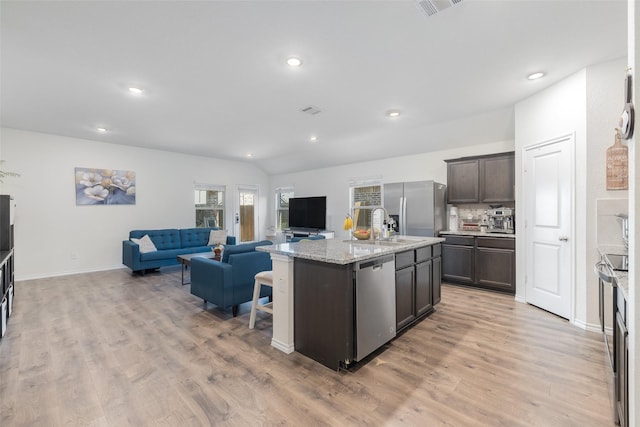 kitchen with appliances with stainless steel finishes, light stone counters, dark brown cabinetry, light hardwood / wood-style floors, and an island with sink