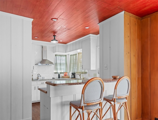 kitchen with wall chimney exhaust hood, wood ceiling, kitchen peninsula, decorative backsplash, and white cabinets