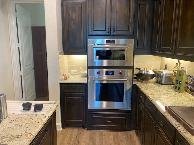 kitchen featuring backsplash, light hardwood / wood-style floors, light stone countertops, dark brown cabinets, and stainless steel double oven