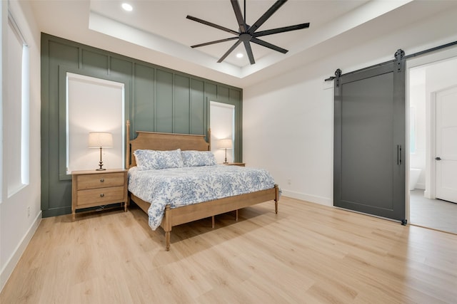 bedroom with ceiling fan, a barn door, light hardwood / wood-style floors, and a tray ceiling