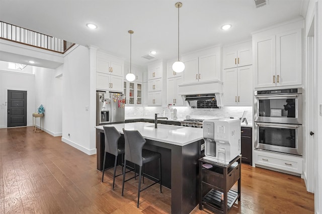 kitchen with stainless steel appliances, an island with sink, white cabinetry, and decorative light fixtures