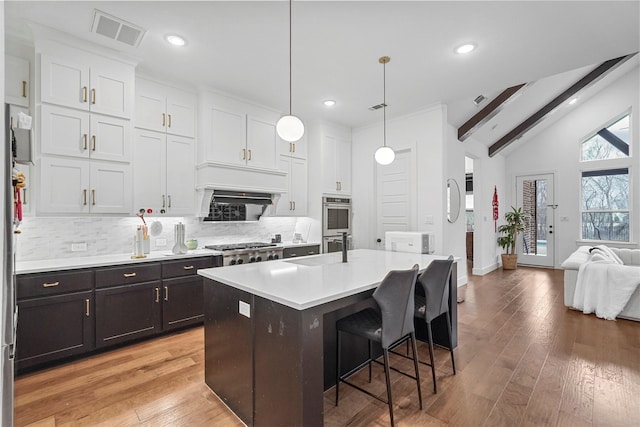 kitchen featuring decorative light fixtures, an island with sink, vaulted ceiling with beams, white cabinets, and backsplash