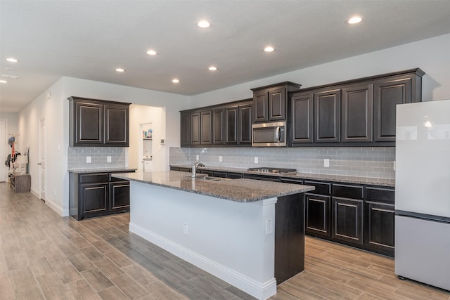 kitchen featuring sink, backsplash, stainless steel appliances, a center island with sink, and dark stone counters