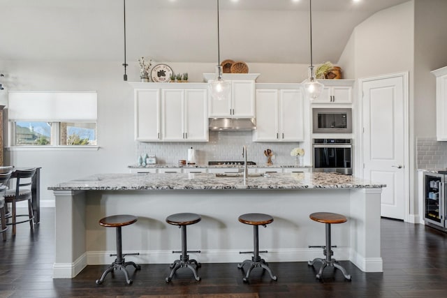 kitchen featuring appliances with stainless steel finishes, decorative light fixtures, white cabinetry, an island with sink, and wine cooler