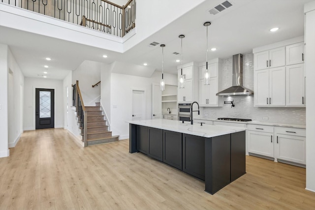 kitchen with wall chimney exhaust hood, gas stovetop, hanging light fixtures, a kitchen island with sink, and white cabinets