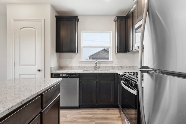 kitchen with sink, stainless steel appliances, dark brown cabinetry, light stone countertops, and light wood-type flooring