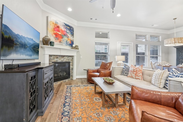 living room with crown molding, ceiling fan, a stone fireplace, and light wood-type flooring