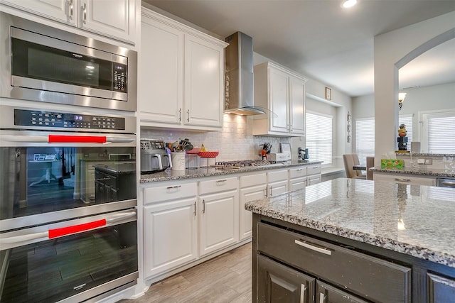 kitchen featuring wall chimney range hood, appliances with stainless steel finishes, white cabinetry, tasteful backsplash, and light stone countertops