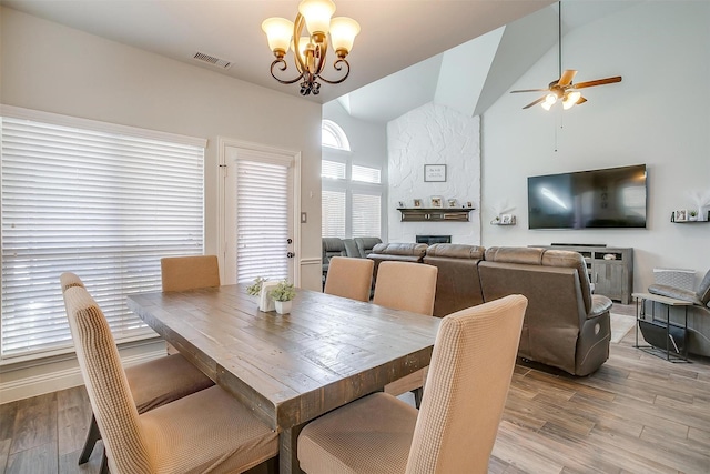 dining area featuring a stone fireplace, ceiling fan with notable chandelier, high vaulted ceiling, and light wood-type flooring