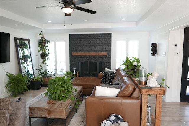 living room featuring a raised ceiling, ceiling fan, light wood-type flooring, a brick fireplace, and recessed lighting