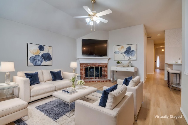 living room featuring ceiling fan, lofted ceiling, a brick fireplace, and light wood-type flooring