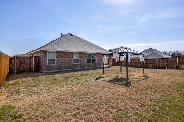 back of house with a fenced backyard, brick siding, a gazebo, a yard, and roof with shingles