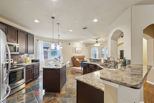 kitchen with dark brown cabinetry, appliances with stainless steel finishes, a center island, light stone countertops, and a sink