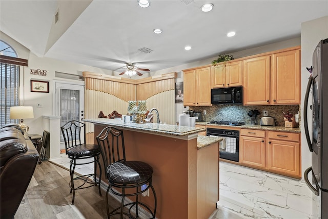 kitchen with tasteful backsplash, a kitchen bar, a kitchen island with sink, black appliances, and light stone countertops