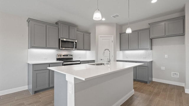 kitchen with gray cabinets, sink, hanging light fixtures, stainless steel appliances, and a center island with sink