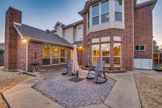 back of house at dusk with a patio area, a shingled roof, a chimney, and brick siding