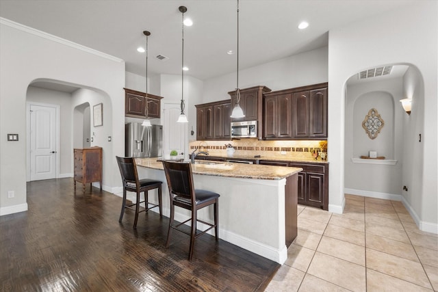 kitchen featuring dark brown cabinetry, tasteful backsplash, hanging light fixtures, stainless steel appliances, and a kitchen island with sink