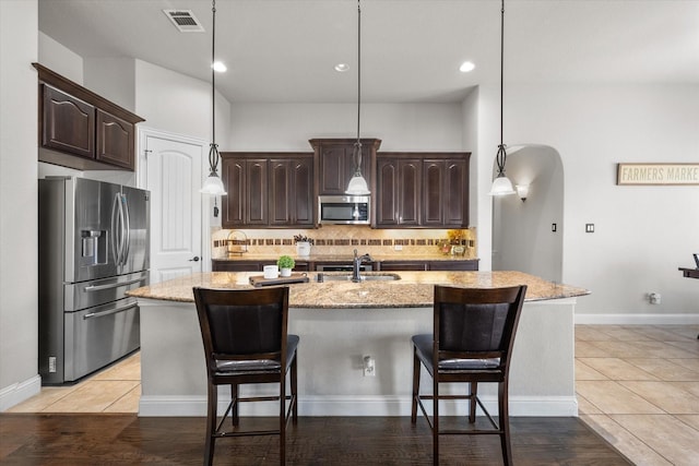 kitchen featuring sink, dark brown cabinets, stainless steel appliances, light stone countertops, and a center island with sink