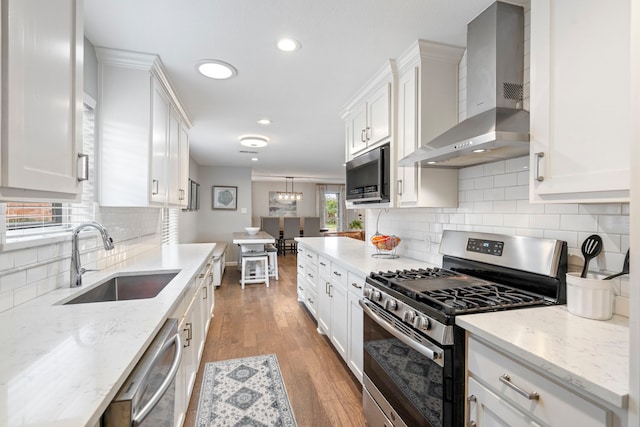 kitchen with wall chimney range hood, sink, appliances with stainless steel finishes, hanging light fixtures, and white cabinets