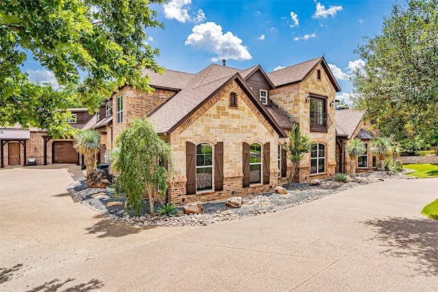 french country style house with stone siding, roof with shingles, concrete driveway, a garage, and brick siding